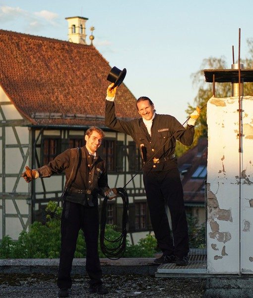 Zwei Schornsteinfeger mit Zylinder vor einem Fachwerkhaus